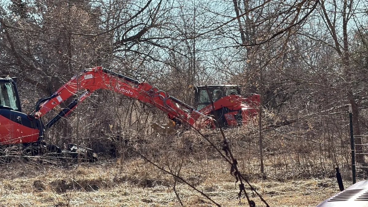Red excavator clearing brush along a fence line, posted by Christopher Gower