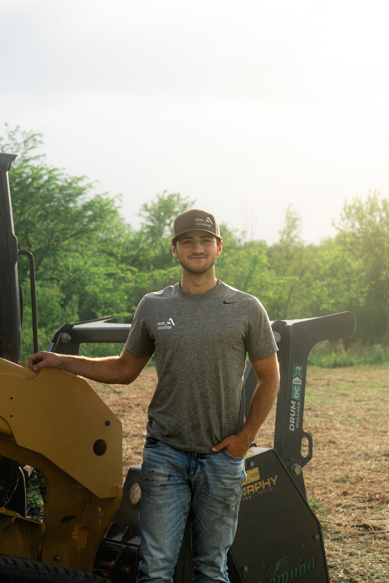 Ashton, owner of Grade A Excavation, smiling beside his CAT skid steer with the mulching head attached