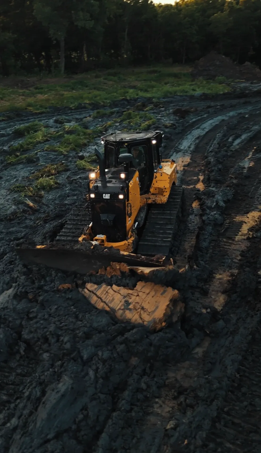 CAT dozer with its lights on pushing logs across muddy ground at dusk